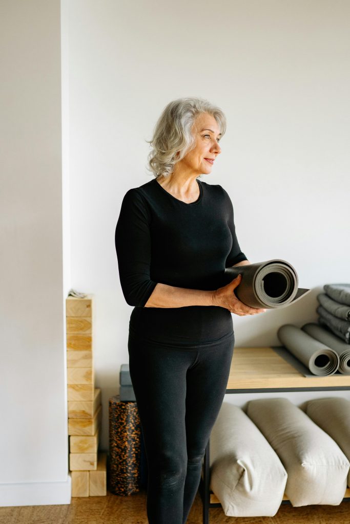 pexels-photo-8437018-8437018 Elderly woman holding a yoga mat, standing in a bright indoor yoga studio.